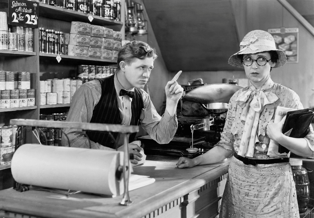Image of an old store in which shoppers had to give a list of products they required to the shopkeeper. The shopkeeper in the image is telling off a female shopper, who looks annoyed. The image represents a time when shopping was full of friction, and modern retail should be moving towards a frictionless checkout environment for better shopper journeys and customer experience